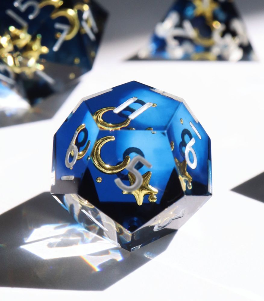 Single Etoile Royale d12 on a white background, more dice are out of focus behind it. The die is a semi-opaque midnight blue with inclusions of yellow stars and moons inside the resin. The numbers on the die are white.