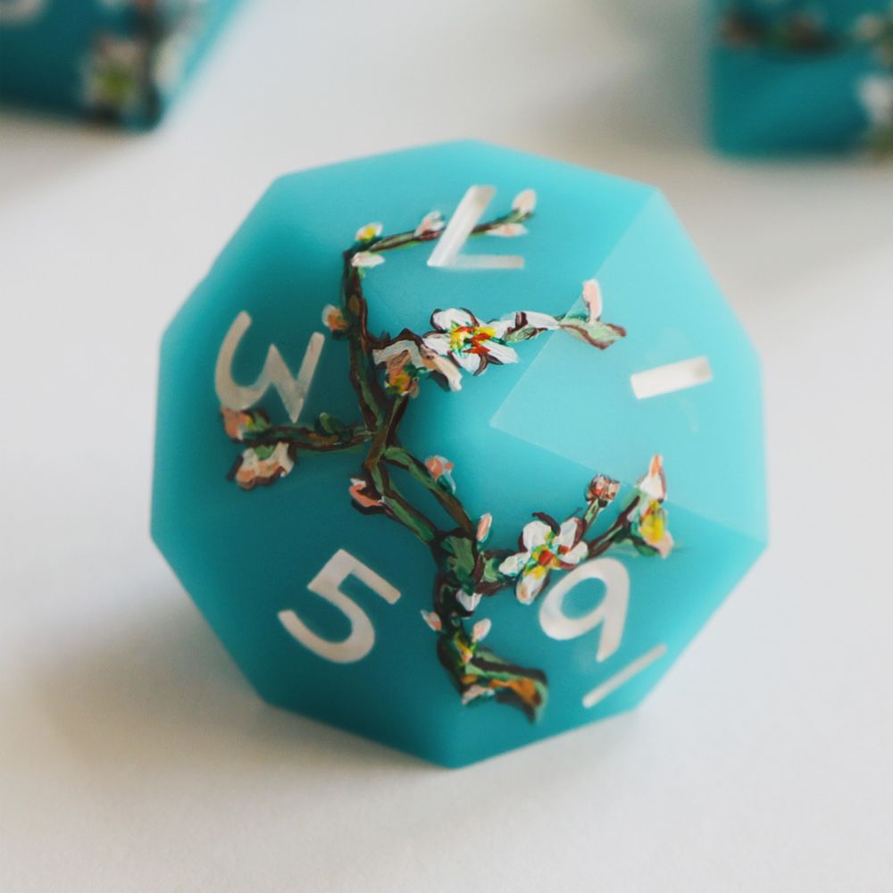 Single light blue matte d10 on a white background. The die has impressionist-style illustrations of white blossoms and multicolored branches painted around it. The numbers on the die are white.