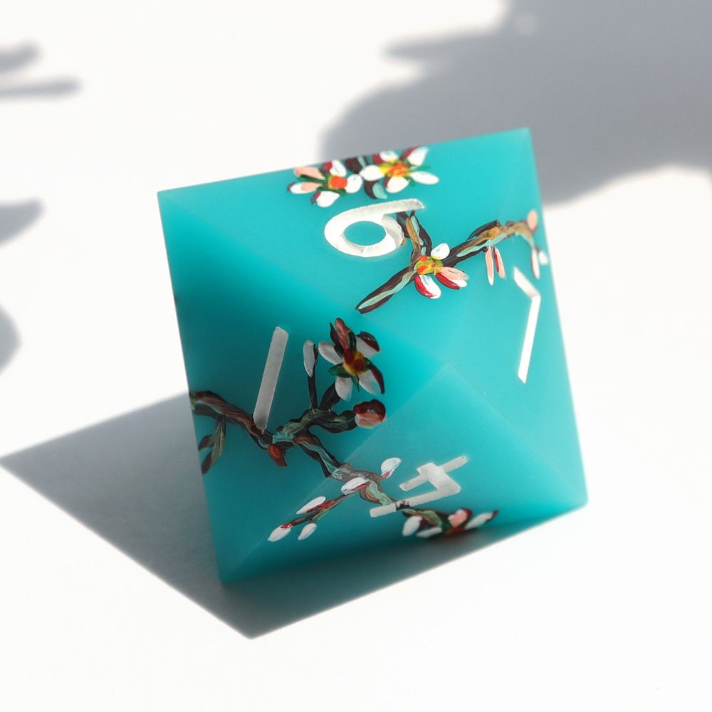 Single light blue matte d8 on a white background. The die has impressionist-style illustrations of white blossoms and green and brown branches painted around it. The numbers on the die are white.