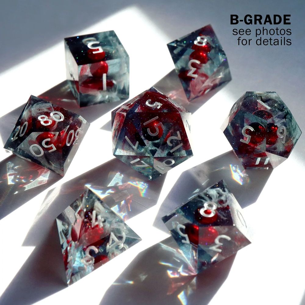 Full set of Ruidus dice arranged in a circle with the D20 in the center. All the dice are a dark grey resin with a red “pearl” in the center representing the moon. Wisps of dark smoke detail is seen. The text reads “B-Grade see photos for details”