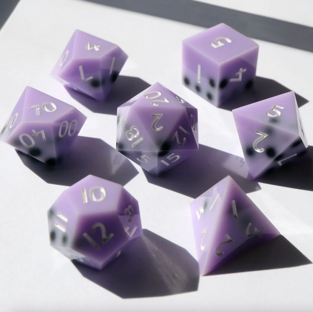 7 piece set of gaming dice arranged in a circle with the D20 in the middle. All the dice are an opaque pastel purple with black "boba pearls" resting on one side of the dice, resembling a boba drink. 