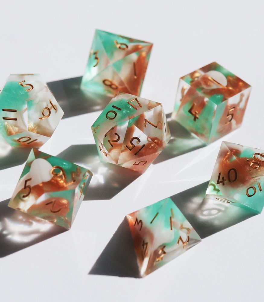 Full set of the Mint Chocolate Chip test set arranged in a circle on a white background with the D20 in the center. All the dice are clear resin with swirls of white, light brown, and mint green resin. The numbers on the dice are gold.