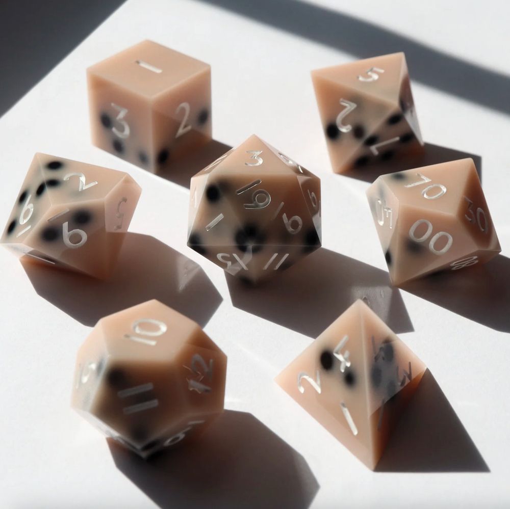 7 piece set of gaming dice arranged in a circle with the D20 in the middle. All the dice are an opaque light brown with black "boba pearls" resting on one side of the dice, resembling a boba drink. 
