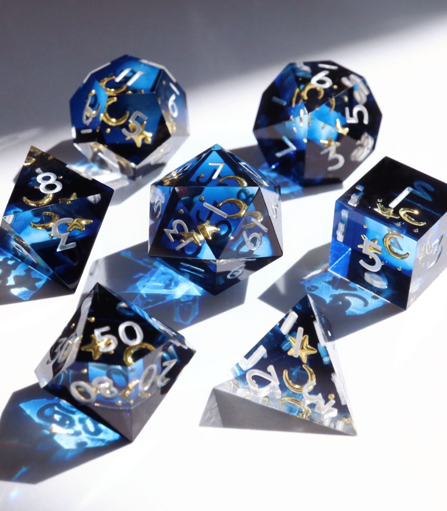 Full 7 piece dice set of Etoile Royale on a white background. The dide are a semi-opaque midnight blue with inclusions of yellow stars and moons inside the resin. The numbers on the dice are white.