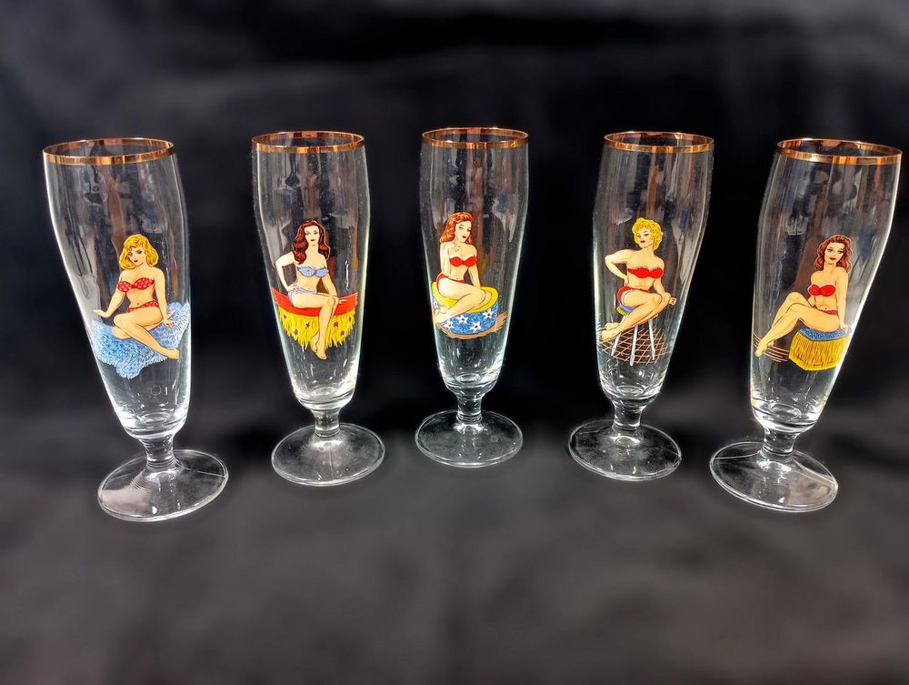 A set of five vintage 1950s peek-a-boo pilsner beer glasses with gold rims and footed bases. Each glass has a different pin-up girl decal in a bikini on the front that disappears to reveal a nude figure on the back. The glasses are in excellent condition.