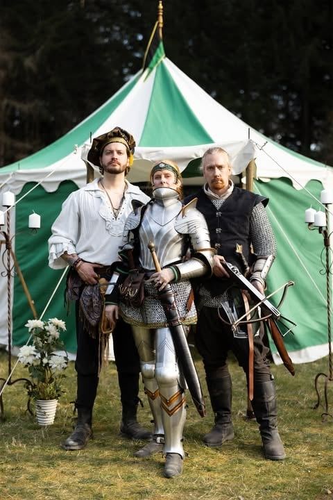A photograph of a woman in full plate armor with two men in medieval clothing standing next to her in front of a green and white pavilion.