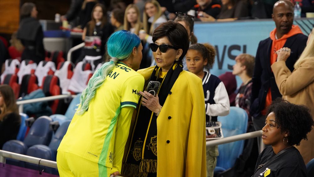 Trinity Rodman and Michele Kang are conversing intently. Michele is wearing her signature yellow cape 
