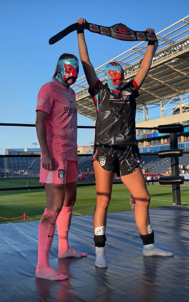 Alyssa Naeher and Sam Staab stand in the middle of a wrestling ring. They’re both wearing their stars uniforms, and a Chicago stars Lucha Libre mask. Sam is holding a wrestling belt over her head. 