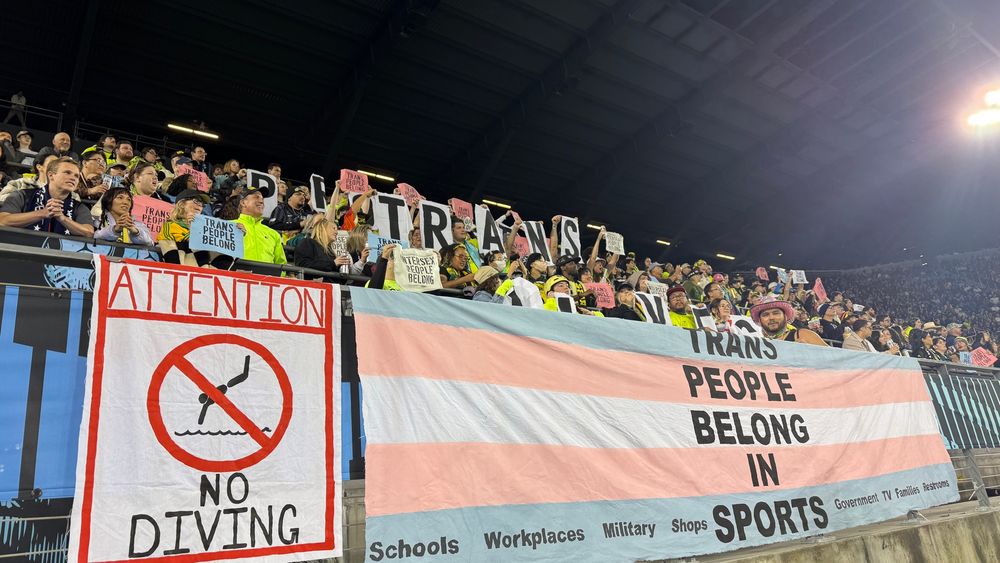 The spirit supporters stand behind a banner that reads “Trans people belong in sports.” Next to the banner is a “no diving” sign 