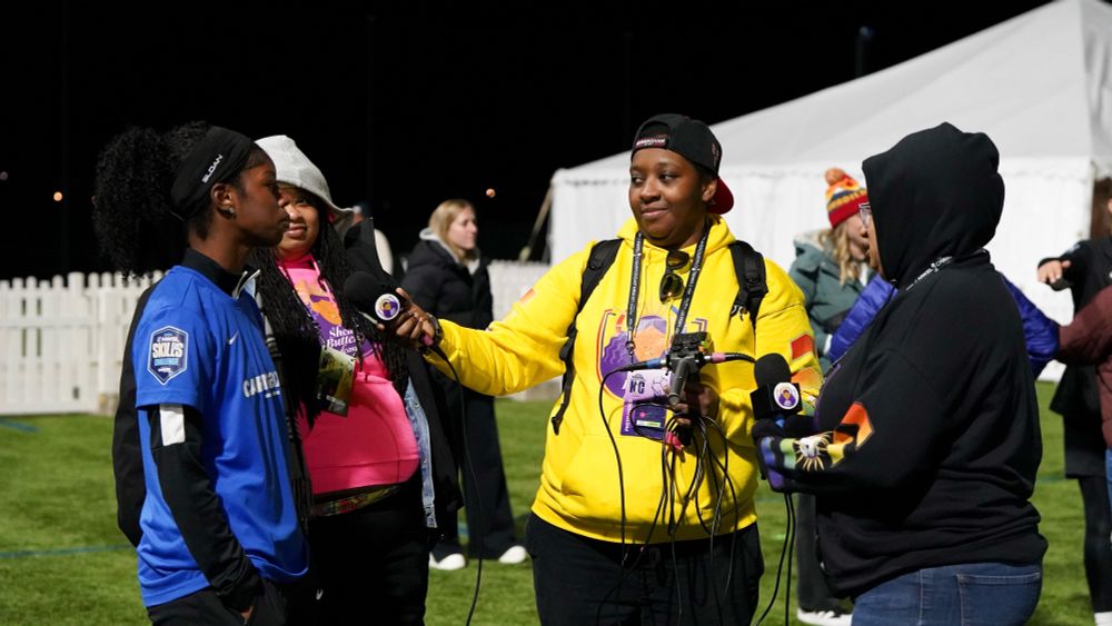 Eboni Christmas is wearing a yellow sweater and holding an audio mixer attached to two microphones. Shes holding a mic out to jameese Joseph, who’s wearing a blue jersey