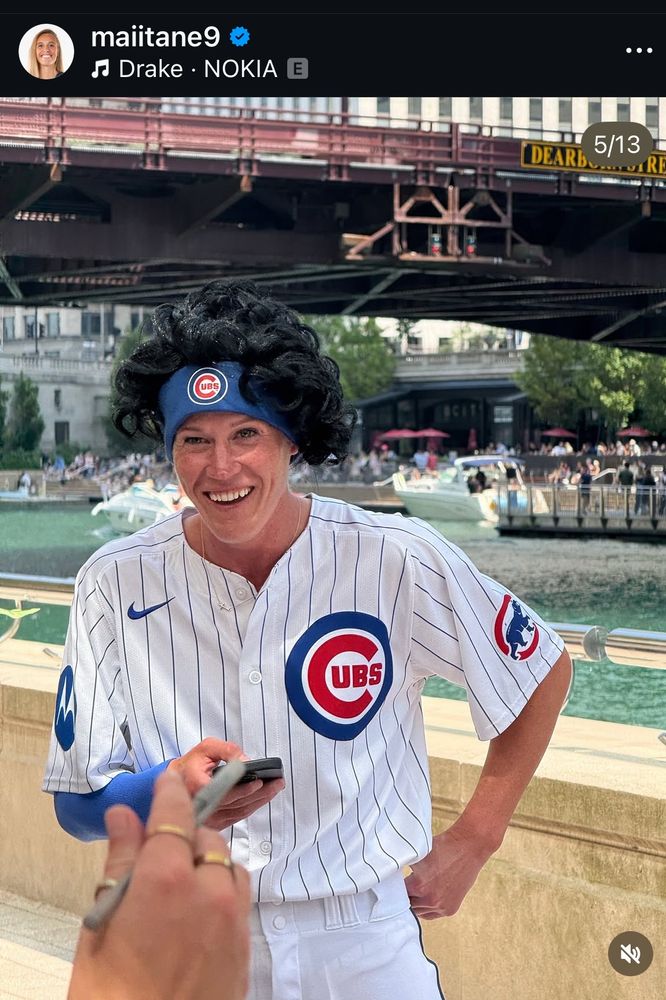 Alyssa is standing against a riverwalk railing wearing a Chicago cups uniform, complete with a black wig and a blue headband 