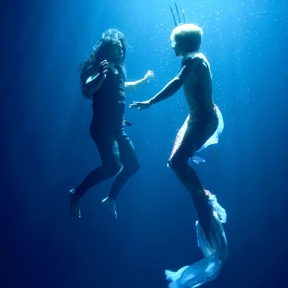 screenshot from ofmd 2x3; stede is appearing as a merperson with a golden tail in ed's gravy basket dream; ed and stede are facing each other, treading water, seen from the side