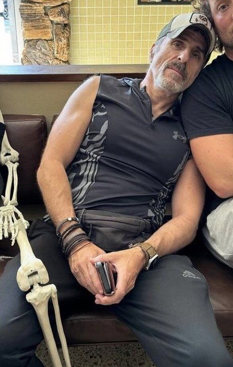 Con O'Neill sitting on a sofa facing the camera, he's wearing a ball cap, gray track pants and what seems to be a sleeveless Lycra shirt in a dark grayish blue. The way the fabric cleaned ngs and stretches emphasizes his tits and slight belly
Borderline obscene