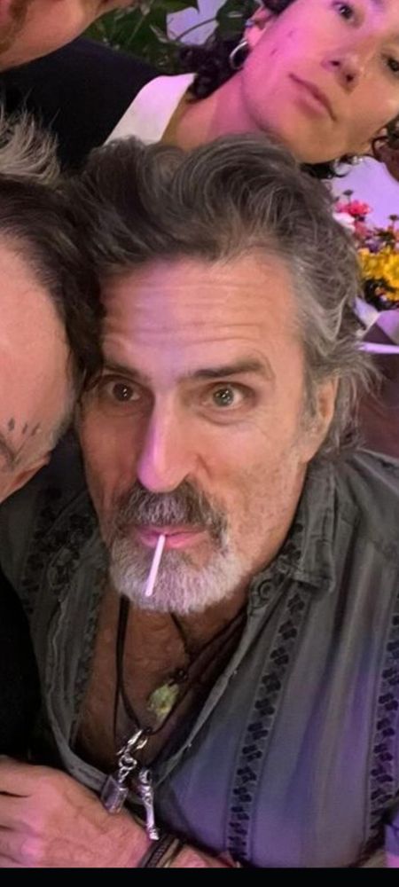 A photo of Con O'neill, shirt partially unbuttoned to expose his ample fluffy chest and necklaces, with a lollipop in his mouth and the most pretty babygirl eyed look  you can imagine like he looks like he's waiting for you to shower him with love and affection and expensive presents