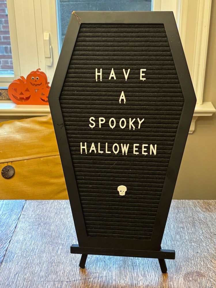 A black coffin shaped letter board with the text “Have a Spooky Halloween”. There is a jack o Lantern shaped candle holder behind it.