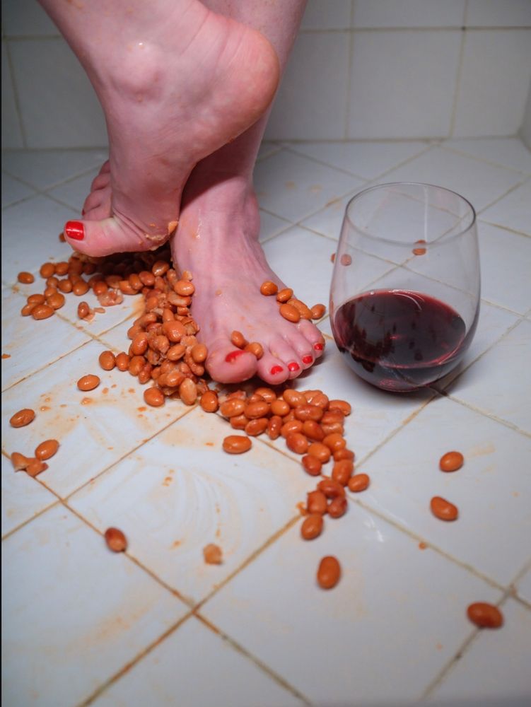 Demure, mindful feet on a white tile floor, surrounded by fava beans, with a glass of red wine in the background.