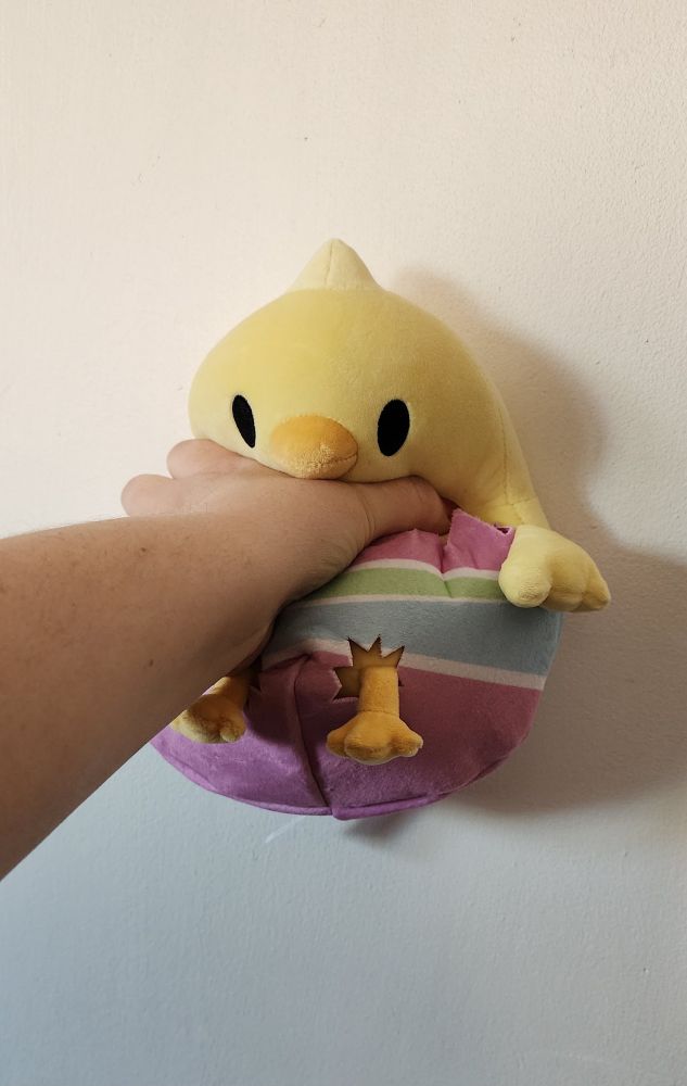 A plushie being held up to and pushed against a white wall by a hand holding it at approximately the neck/abdomen area. The plushie is of a yellow bird with black embroidered eyes, and it's lower half in a cracked half of a decorated eggshell of pink, green, blue, and pink again stripes separated by white stripes. Little bird legs hang out the bottom of the eggshell as if they broke through, and small wings hang out just above the half of the eggshell.