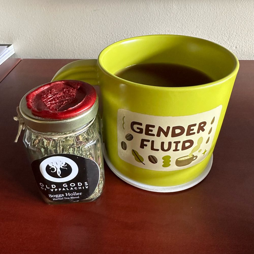 a green mug sitting on a desk with the words “gender fluid” on the front from the target pride collection. next to it is a small glass jar of and herbal tea called “Boggs Holler” herbal tea blend from Old Growth Alchemy. there’s a label on the jar with the Old Gods of Appalachia logo and a wax seal on top of the jar’s lid.