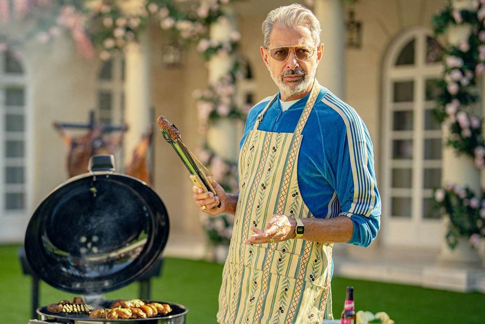 Jeff Goldblum on the set of the Netflix series Kaos where he plays the role of Zeus. He's wearing an apron and standing by a grill outside of a beautiful mansion. 