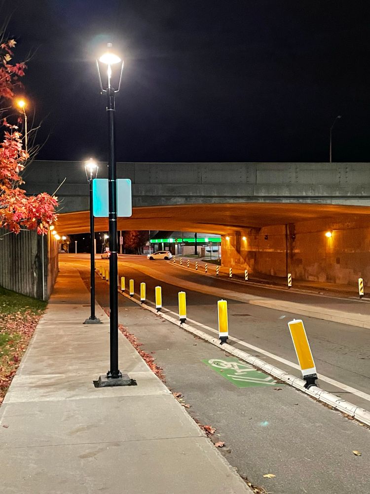 Photo of two lamp posts in the middle of a sidewalk next to a cycling facility and a car lane.  