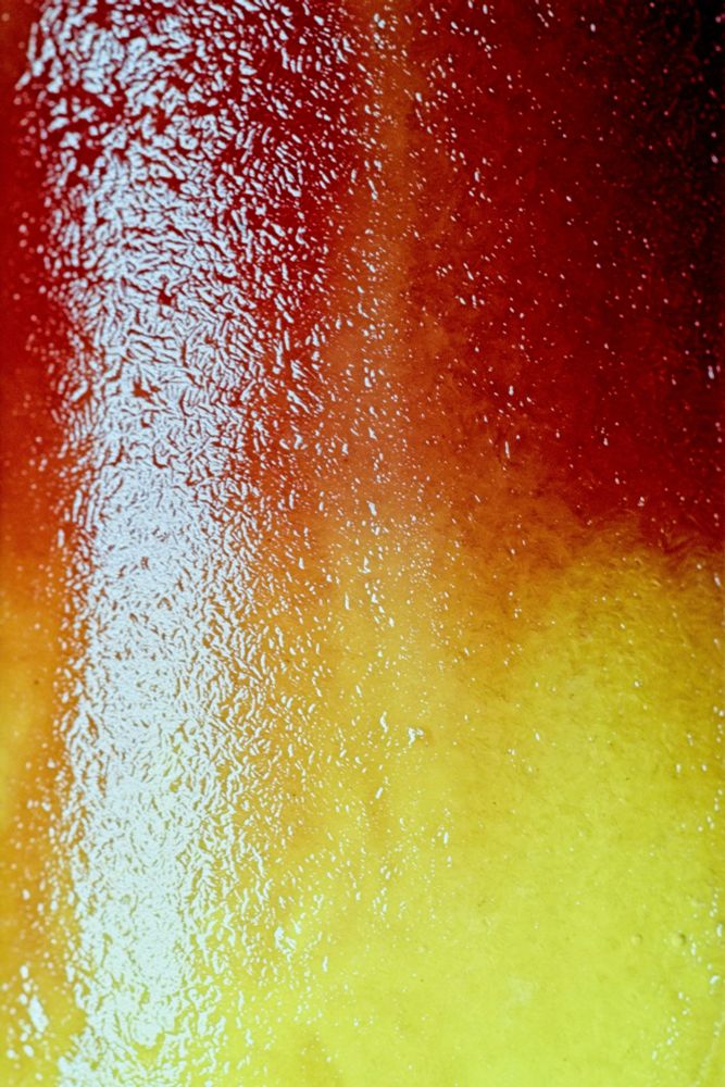 This image captures the interplay between smooth and textured surfaces. The composition shows a gradient from deep red at the top flowing down through warm orange tones to bright yellow at the bottom. The left side of the image features a heavily textured area with countless tiny water droplets creating an intricate, almost lace-like pattern of light and shadow. This textured zone contrasts dramatically with the right side, where the surface appears completely smooth with unbroken color transitions. The lighting creates a luminous quality, making the colors appear to glow from within while the water droplets add a tactile, almost touchable quality to the surface.