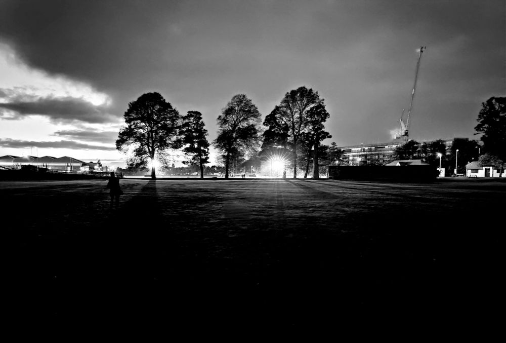 A black and white photograph captured at twilight showing an urban landscape dominated by artificial glow. The foreground is a large, dark open field with a solitary silhouetted figure standing in the vast empty space. Across the middle distance, a row of mature trees creates a dark barrier, behind which multiple street lamps and sports lights create brilliant star-burst effects - the urban glow piercing through the dusk. A construction crane rises on the right, and low buildings line the horizon beneath dramatic clouds where natural light is fading. The image captures that threshold moment when the city's artificial illumination overtakes the last natural light, creating a complex interplay between darkness and urban radiance.