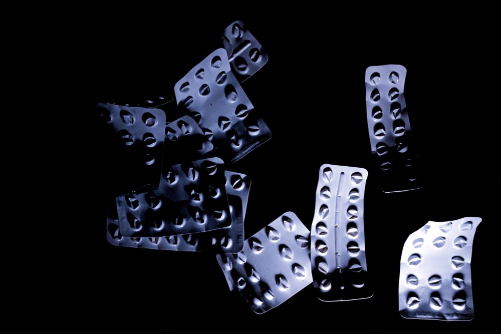 This black and white photograph shows empty medicine blister packs scattered across a dark background. The metallic strips, which once held pills or tablets, are now hollow with their contents removed, creating patterns of oval and circular voids. The strips overlap and curl naturally, some catching dramatic lighting that creates strong shadows and highlights against the deep black surface. The arrangement appears organic and unplanned, as if the empty packets have accumulated over time. The clinical, silvery appearance of the medical packaging contrasts starkly with the darkness surrounding them, while the empty spaces where medication once sat tell a story of treatment completed and progress made along a road to healing, may-be...