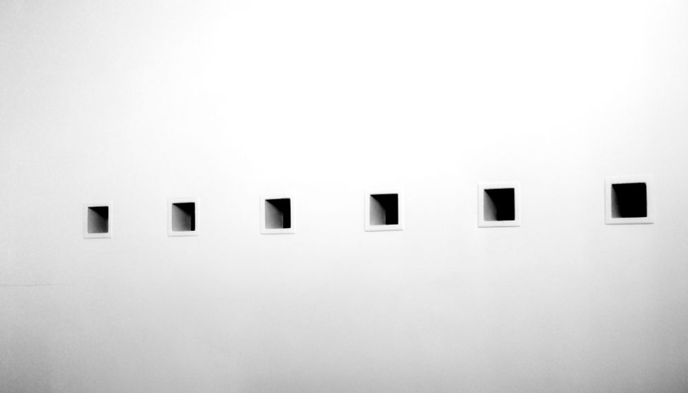 A minimalist architectural photograph showing six square recessed windows arranged in a horizontal line across a pale wall. The windows graduate in size from small on the left to large on the right, creating a rhythmic progression. Each opening reveals a dark interior that contrasts sharply with the light-colored wall, while subtle gradients of shadow transition from the bright frames into the black depths.