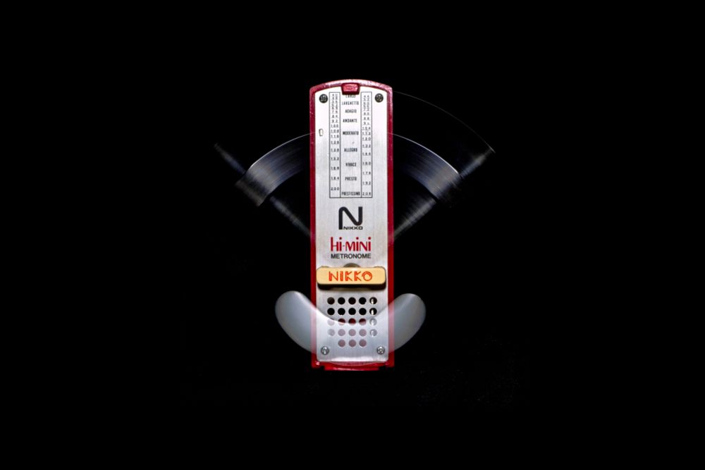 A red metronome photographed against a pure black background. The metronome's pendulum arm is captured in motion, creating symmetrical motion blur arcs that demonstrate the alternating movement between left and right positions. These translucent, ghostly trails extend horizontally from the central mechanism, visually representing the alternating rhythm that defines the metronome's function. The metronome body remains sharp and stationary in the center, showing clear tempo markings, while the blurred pendulum traces make the alternating motion visible as ethereal streaks frozen in time.