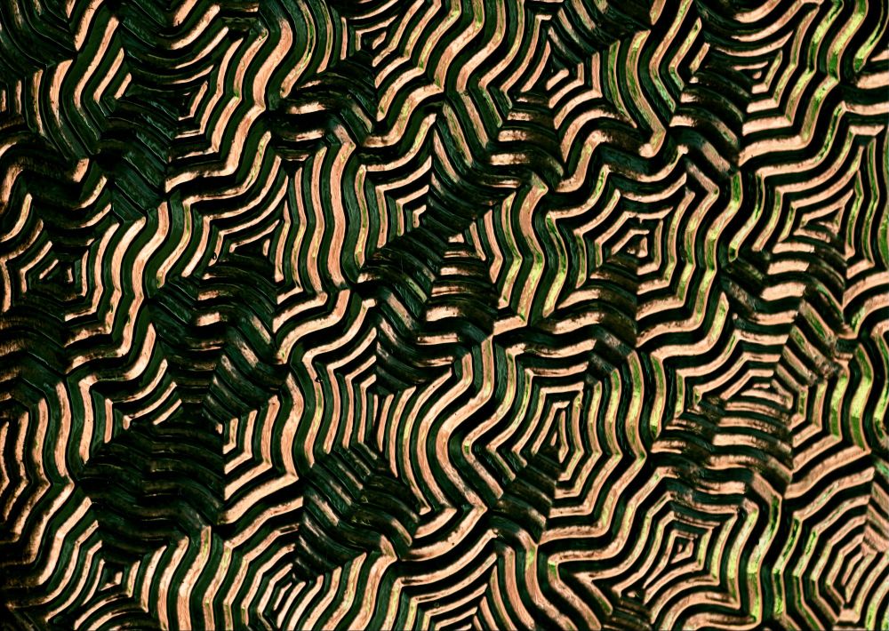 An abstract photograph displaying complex geometric patterns with concentric bands and angular zigzag formations in rich earth tones of deep green, bronze, cream, and metallic highlights. The image features flowing geometric wave structures that create rhythmic, tessellated patterns across the frame. Each colored band follows precise angular paths that fold and intersect, creating a mesmerizing optical geometry reminiscent of topographical contour lines or crystalline lattice structures. The metallic highlights emphasize the geometric precision of these undulating, chevron-like formations that seem to ripple in mathematical harmony across the composition, with the warmer color palette giving it an organic, almost wood-grain quality.