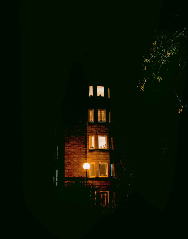 A narrow multi-story brick building photographed at night, surrounded by complete darkness. Four levels of windows glow with warm yellow light from inside, arranged vertically up the red-brown brick facade. A single streetlamp illuminates the lower left corner. The building appears isolated and suspended in a black void, with only a fragment of tree foliage barely visible in the upper right catching ambient light.
