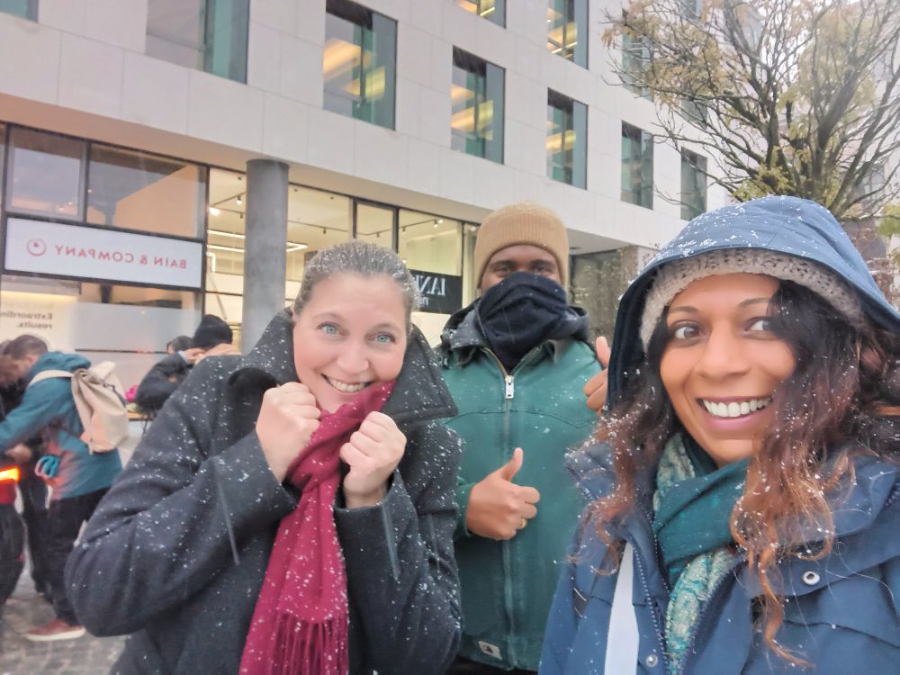 Photo of Ele, Shomari and Saher, wrapped up in winter coats, standing outside. They are covered in a light dusting of snow.