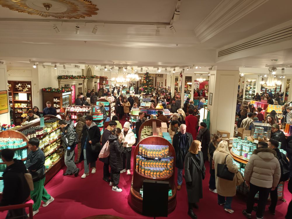 Photo of an inside of a shop with lots of people crowding round Christmas gifts, tea, coffee, chocolates, biscuits. Lots of Christmas trees are visible. Interior of Fortnum and Masons, London