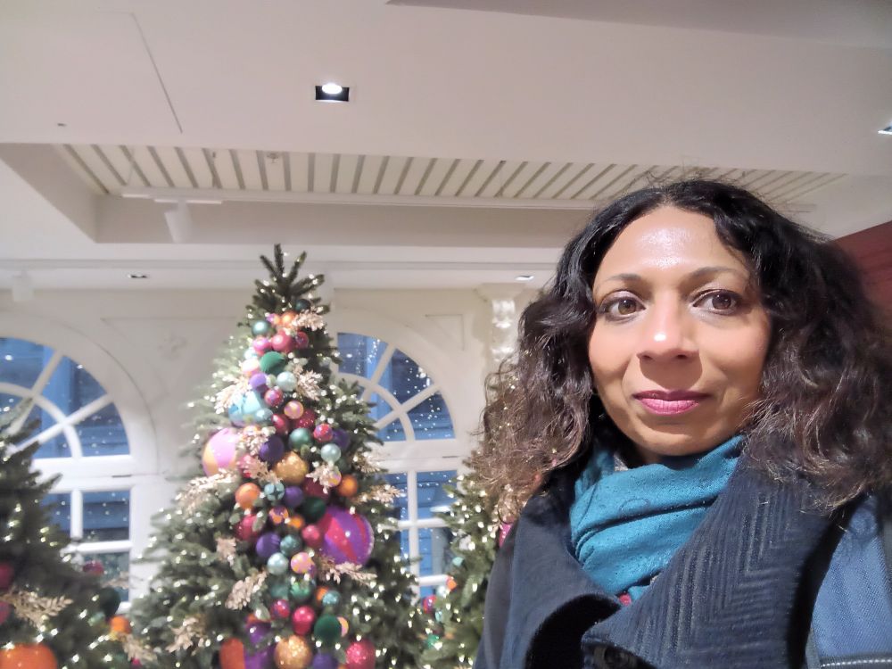 Selfie of me smiling to the camera,  next to brightly decorated Christmas trees. Interior of Fortnum and Masons in London 