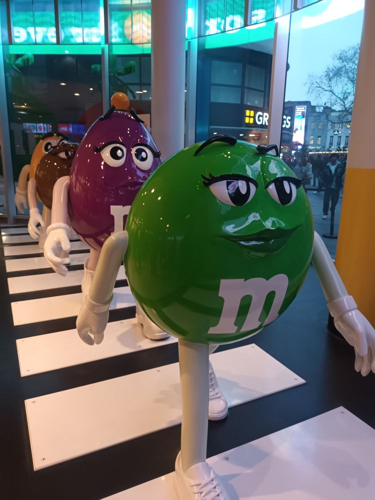 Picture of life sized M&Ms walking happily.  They are green, purple,  brown and yellow 