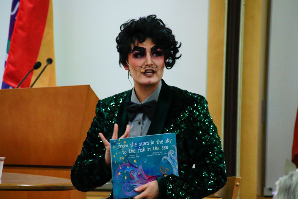 Drag king Cyril Cinder speaking at a conference. He is holding a copy of the picture book "From the Stars in the Sky to the Fish in the Sea" by Kai Cheng Tom.