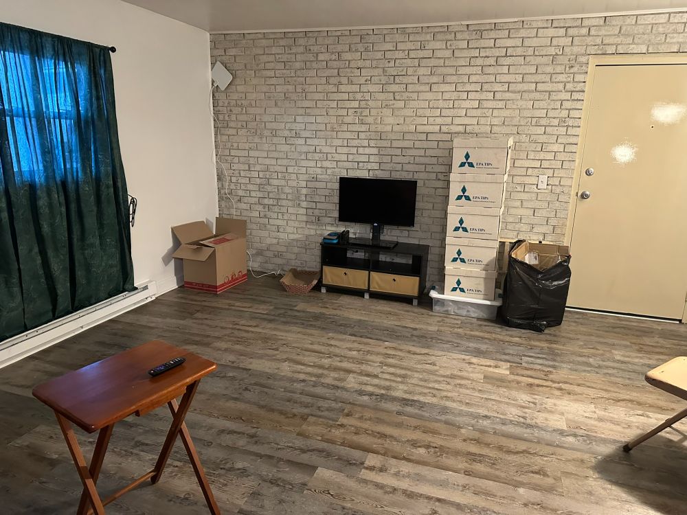 A view of the sparsely-decorated living room. The far wall, from left to right, has an open cardboard box, a cardboard scratching pad for a cat, a medium-sized TV set on a stand, five paperwork boxes stacked atop a short, wide clear plastic tote, and an black open bag of trash. The far wall is made of brick and painted white.