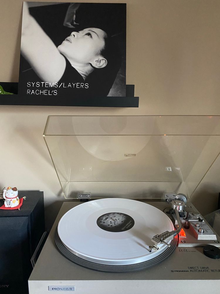 White vinyl record spins on a turntable. The cover of the record sits above on a shelf, which depicts a person looking up in black and white. The title “Systems/Layers” and artist’s name “Rachel’s” is on the bottom lower left corner of the cover in white text.
