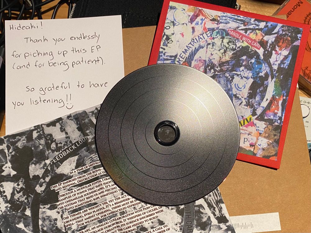 A black CD sits in the middle of the table surrounded by a paper CD cover with a red border, black-and-white, fold-out paper that gives background of the album, and a handwritten note that reads “Hideaki! Thank you endlessly for picking up this EP (and for being patient). So grateful to have you listening!!”