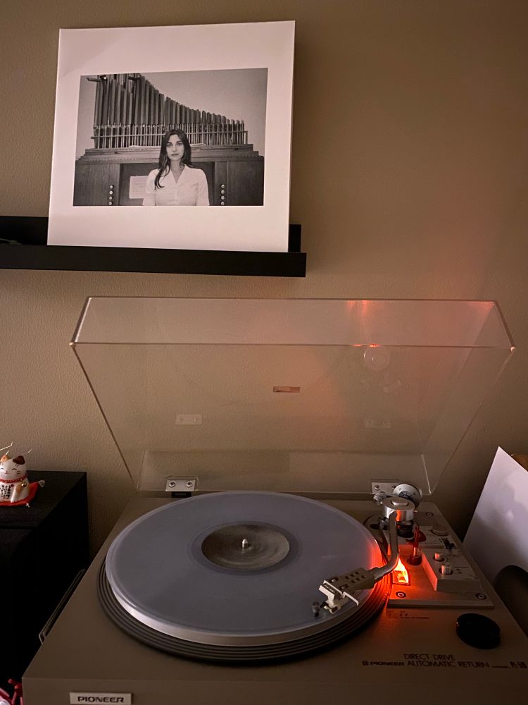 Record player spinning a clear/white vinyl record. Above is the white cover of “Sacrificial Code” by Kali Malone; a black and white picture of her sitting in front of an organ is at the center of the cover.
