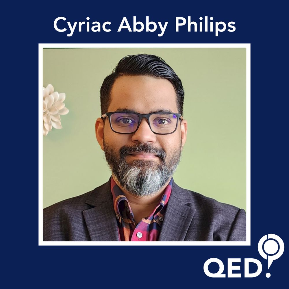 Cyriac Abby Philips - a brown man with short brown hair, a smart beard and glasses. He's wearing a suit jacket and a multicoloured shirt. 