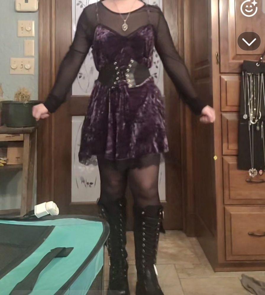 Woman wearing sheer tights, sheer high neck longsleeve under purple velvet babydoll dress with lace trim. She's wearing knee high combat boots, a corset belt, and a silver necklace.