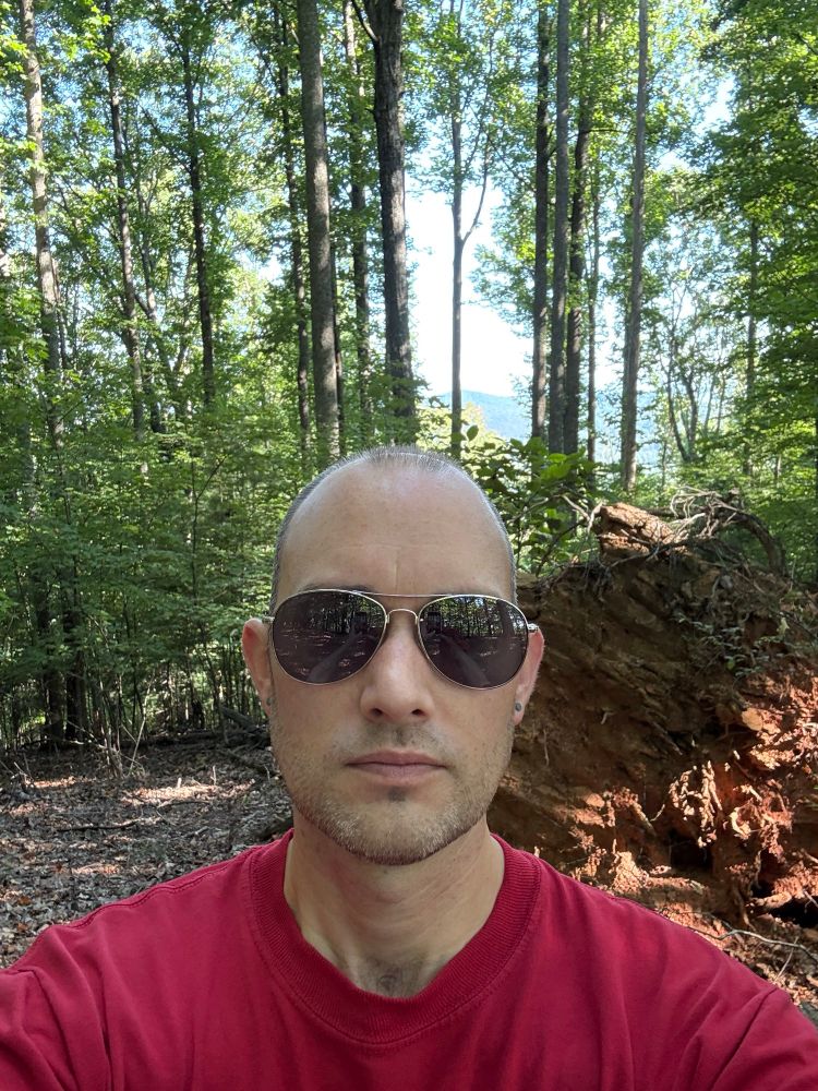 Vance standing in front of a downed tree and ridge-line mountain view in the background. 