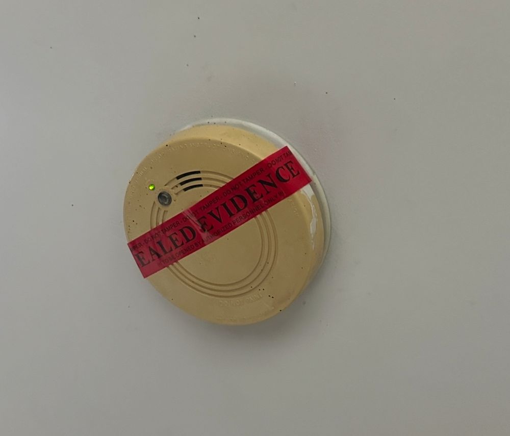 Photo of a smoke detector on a ceiling. There’s red tape across it that says “SEALED EVIDENCE”