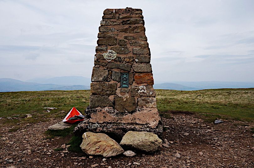Stone built trig point sits on bare ground but the rest of the fell top is grassy. An overcast day but some fells just visible in the distance. At the foot of the trig point is a plastic(?) triangle of red about a foot across.