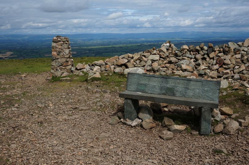 A stone built trig point sits on rocky ground with a stone wall behind and a stone built bench in the foreground.