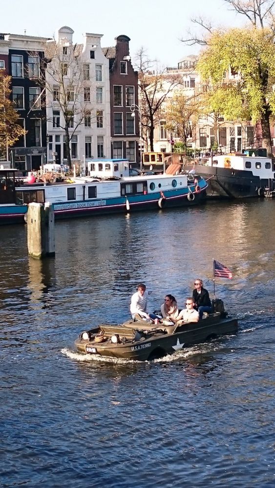 Photograph of an ex military amphibious truck, known as a DUKW, motoring along a canal in the Netherlands.
On the truck are four people enjoying the sunshine as they travel through the water. In the background are tall Dutch buildings and some barges.