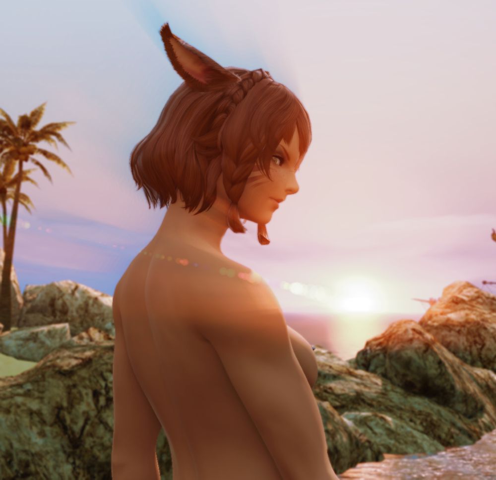 M'ahra stood on the beach at sunset with her back to the camera. She's topless and looking back over her shoulder toward the camera.