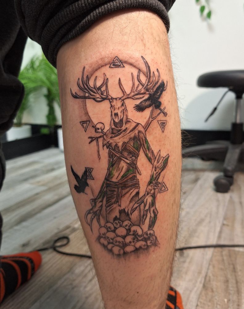 A tattoo on the right calf leg of a leshen from The Witcher 3, mostly black and white, with some green shading for the moss. The Witcher signs are places surrounding the leshen, and the leshen itself is above a pile of human skulls. Two crows are mid flight, one on each side of the leshen.