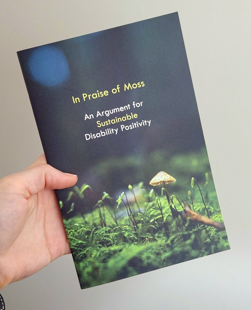 A zine held in someone's hand. The cover features a close-up of moss and reads "In Praise Of Moss - An Argument for Sustainable Disability Positivity" 